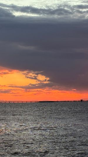 Vibrant coastal sunset over rippling water with a silhouetted bridge on the horizon, glowing orange sun and dramatic purple clouds