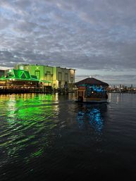 Evening marina scene with a green-lit waterfront restaurant casting neon reflections on rippling water and a thatched-roof tiki boat glowing blue as it cruises under a cloudy sky.
