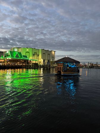 Evening marina scene with a green-lit waterfront restaurant casting neon reflections on rippling water and a thatched-roof tiki boat glowing blue as it cruises under a cloudy sky.