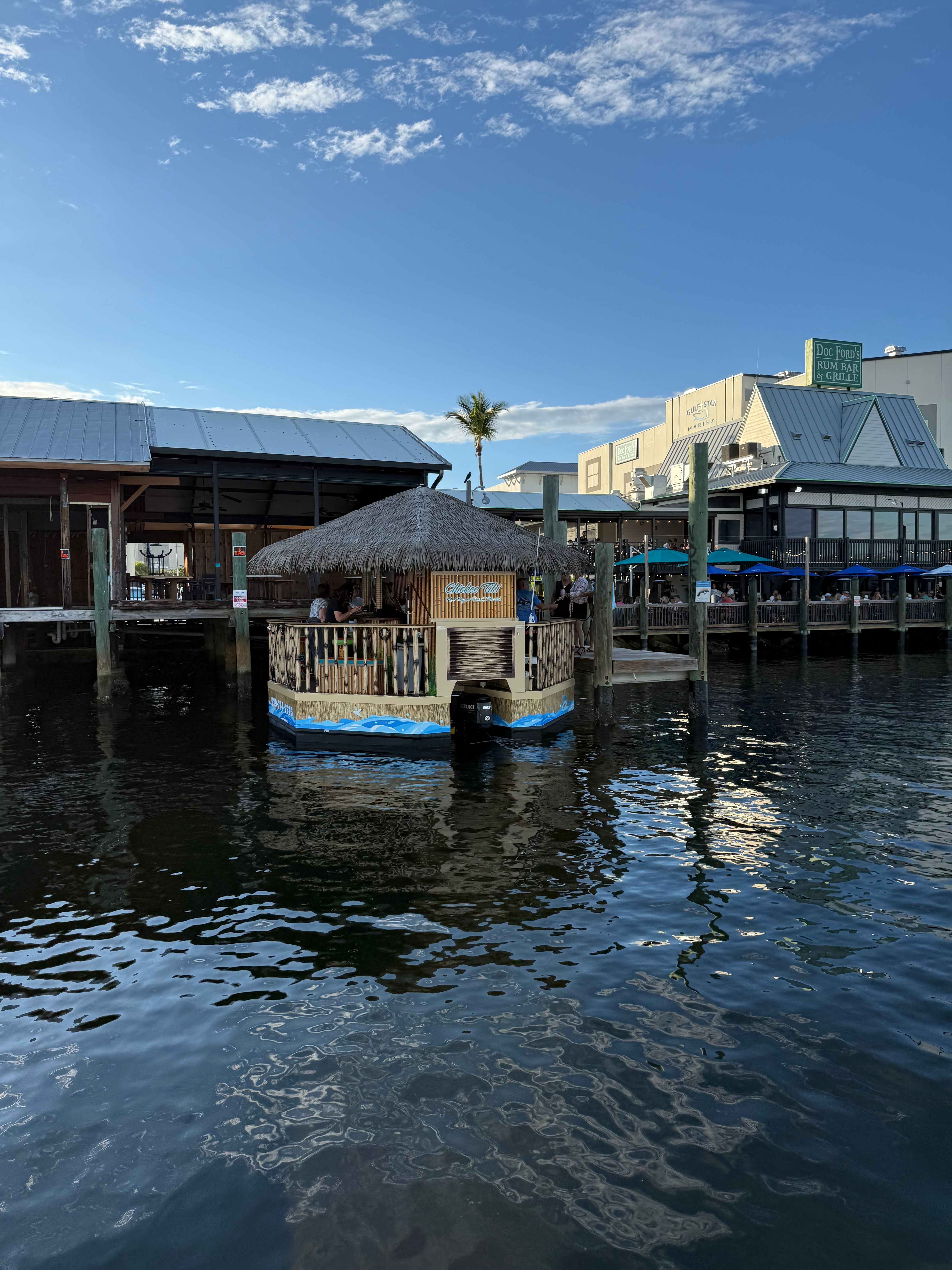 Floating tiki-bar with a thatched roof docked at a sunny marina, palm tree and waterfront restaurants reflecting on calm blue water