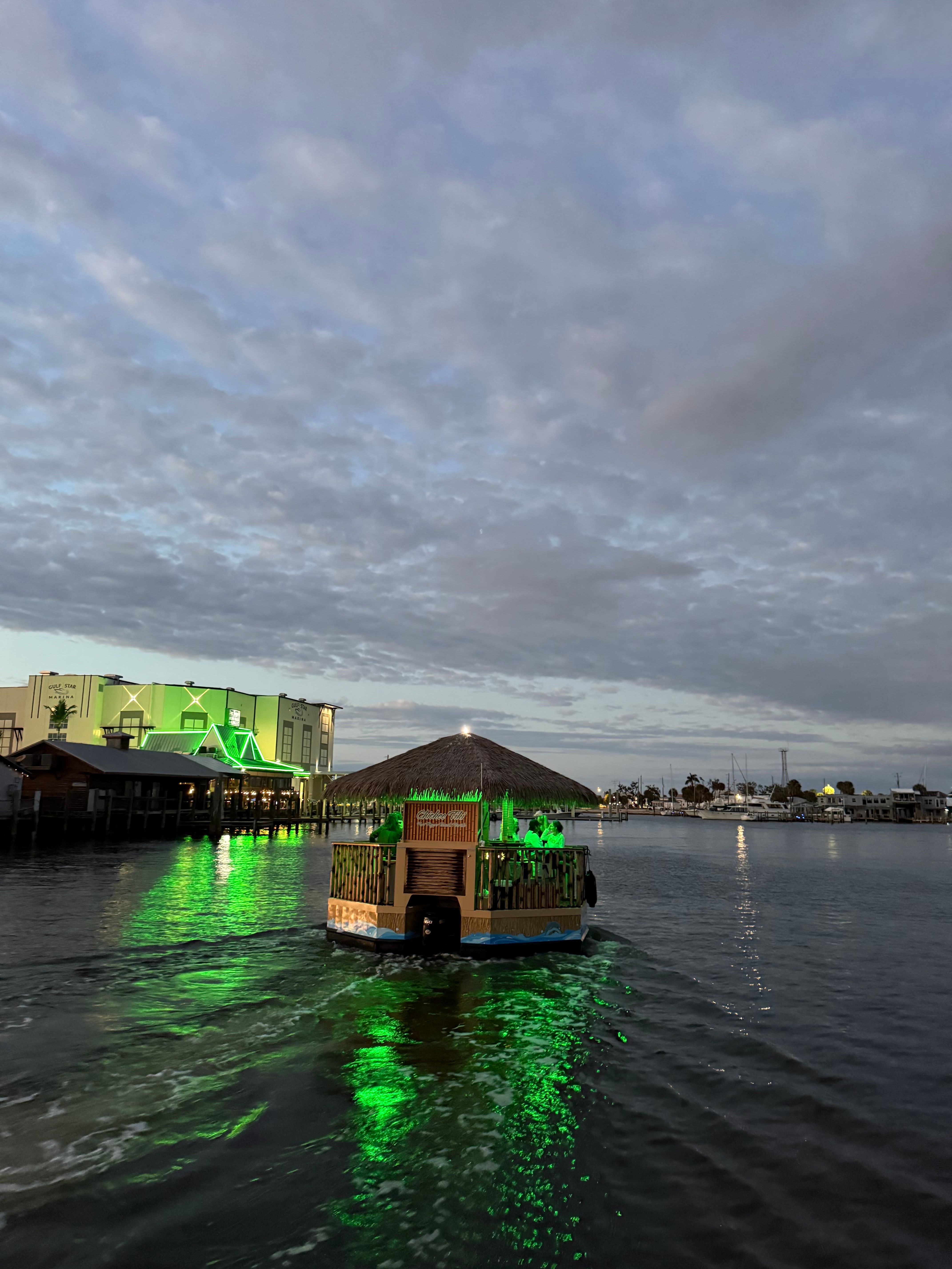 Thatched-roof tiki boat cruising a coastal marina at dusk, green lights reflecting on calm harbor water under a cloudy sky.