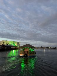 Thatched-roof tiki boat cruising a coastal marina at dusk, green lights reflecting on calm harbor water under a cloudy sky.