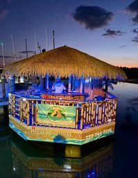 Floating tiki bar with a large thatched roof moored at a marina at dusk, blue and warm lights illuminating bamboo railings and a person behind the bar, calm water reflecting the colorful glow.