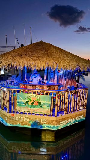 Floating tiki bar with a large thatched roof moored at a marina at dusk, blue and warm lights illuminating bamboo railings and a person behind the bar, calm water reflecting the colorful glow.