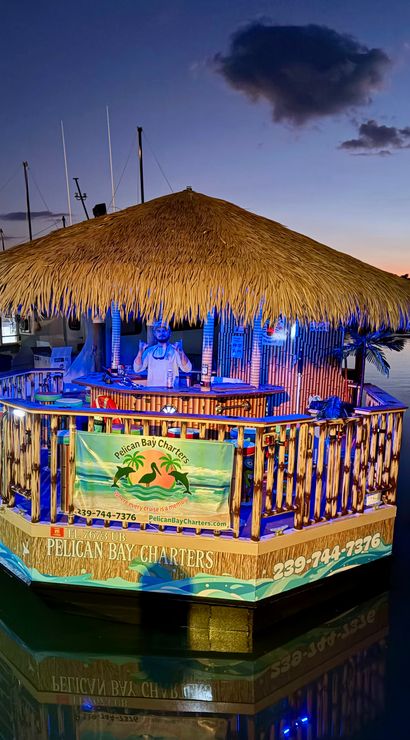 Floating tiki bar with a large thatched roof moored at a marina at dusk, blue and warm lights illuminating bamboo railings and a person behind the bar, calm water reflecting the colorful glow.