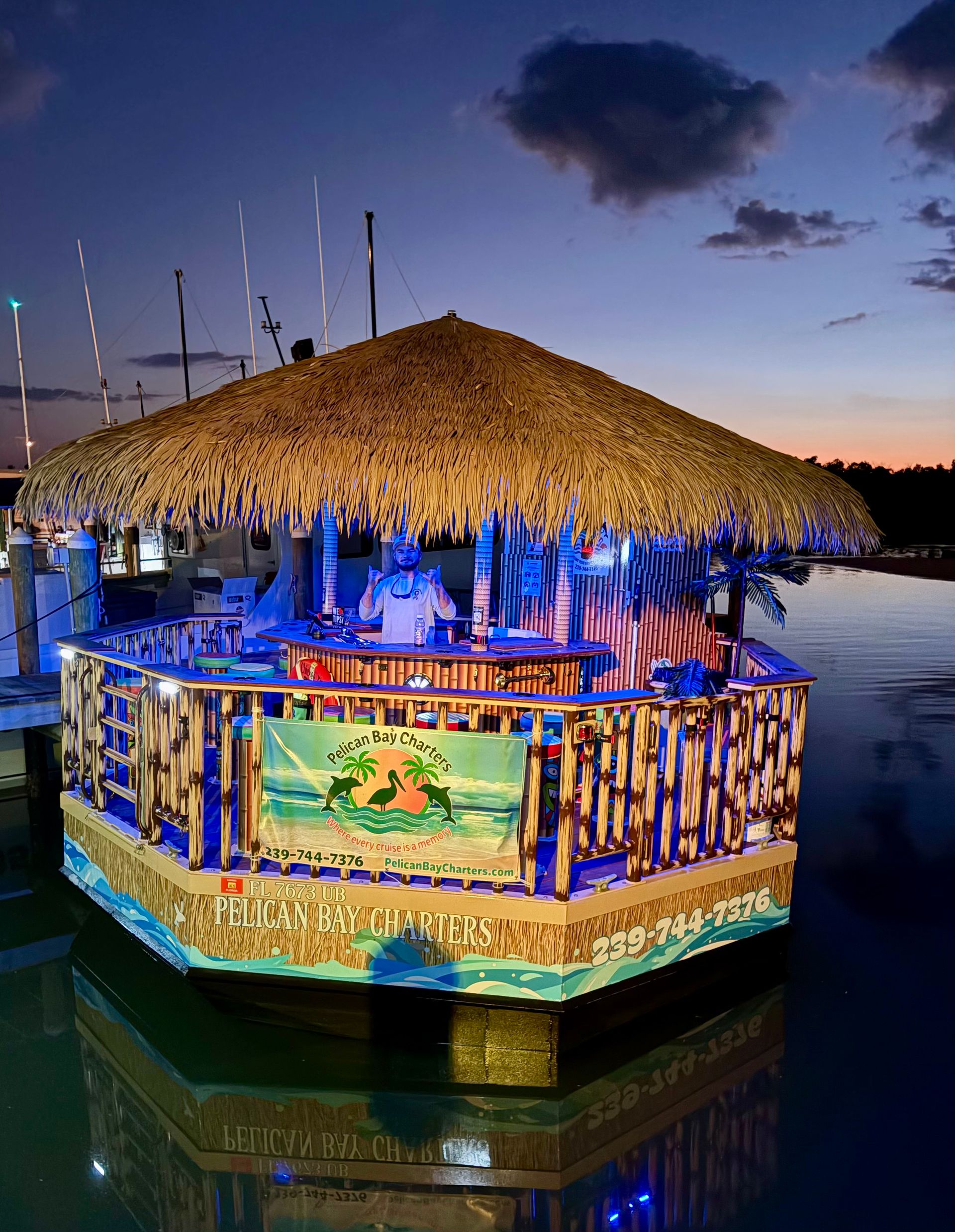 Floating tiki bar with a large thatched roof moored at a marina at dusk, blue and warm lights illuminating bamboo railings and a person behind the bar, calm water reflecting the colorful glow.