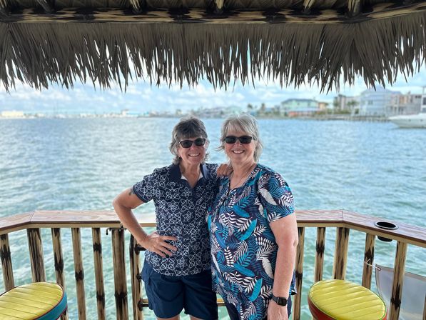 Two smiling women in sunglasses on a tiki-style waterfront deck overlooking a turquoise bay with coastal homes in the distance