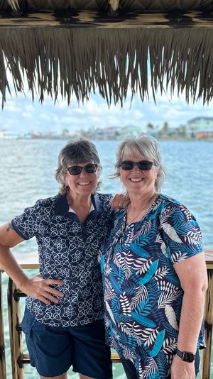 Two smiling women in sunglasses on a tiki-style waterfront deck overlooking a turquoise bay with coastal homes in the distance