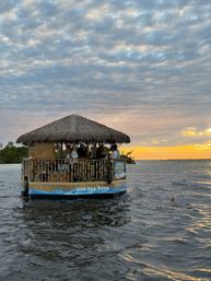 Thatched-roof tiki boat cruising coastal waters at sunset, passengers silhouetted against a golden horizon and dramatic cloud-filled sky.