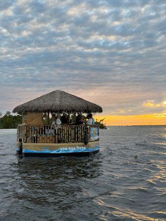 Thatched-roof tiki boat cruising coastal waters at sunset, passengers silhouetted against a golden horizon and dramatic cloud-filled sky.