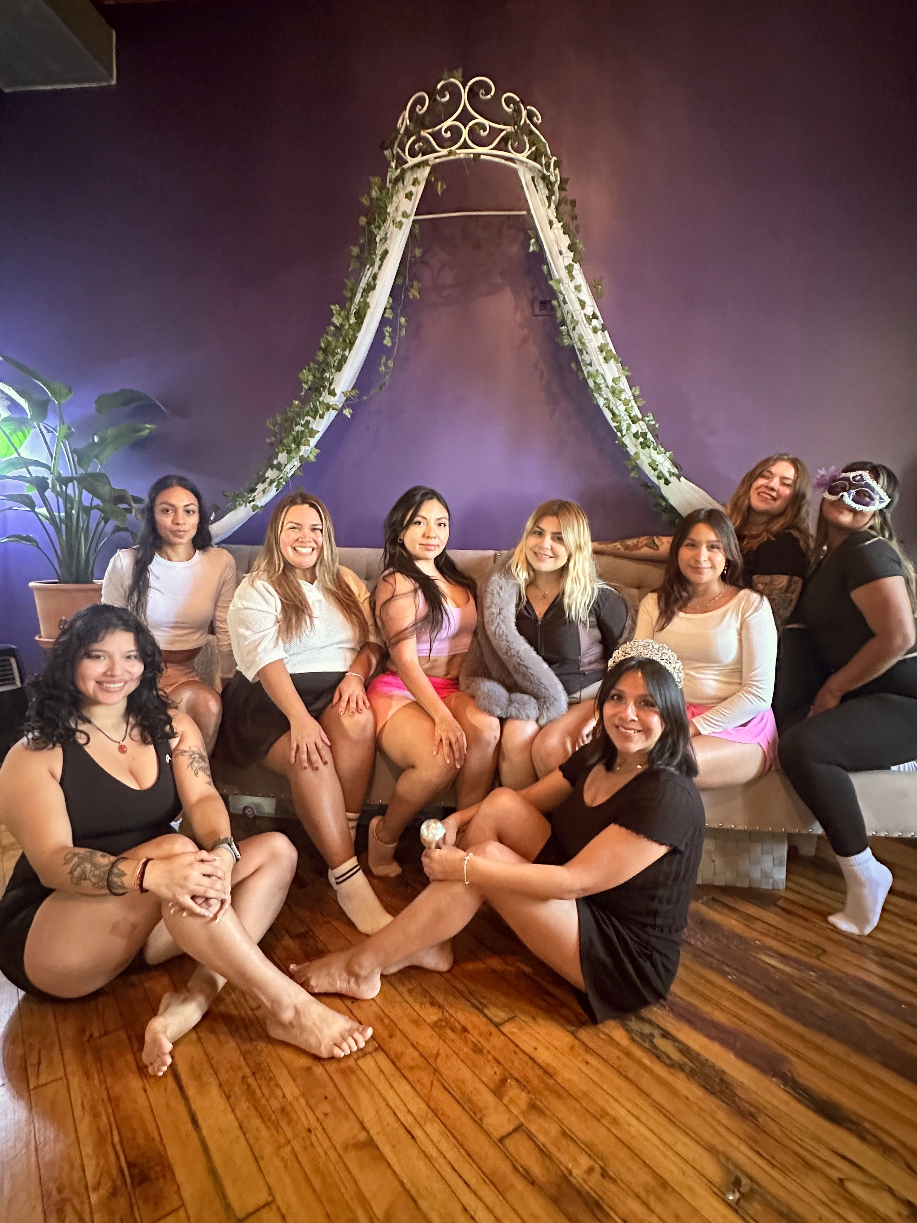 Nine women smiling in a cozy purple-walled lounge with hardwood floors, posing under an ivy-draped canopy; one wears a tiara for a bachelorette-style celebration.