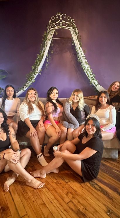 Nine women smiling in a cozy purple-walled lounge with hardwood floors, posing under an ivy-draped canopy; one wears a tiara for a bachelorette-style celebration.