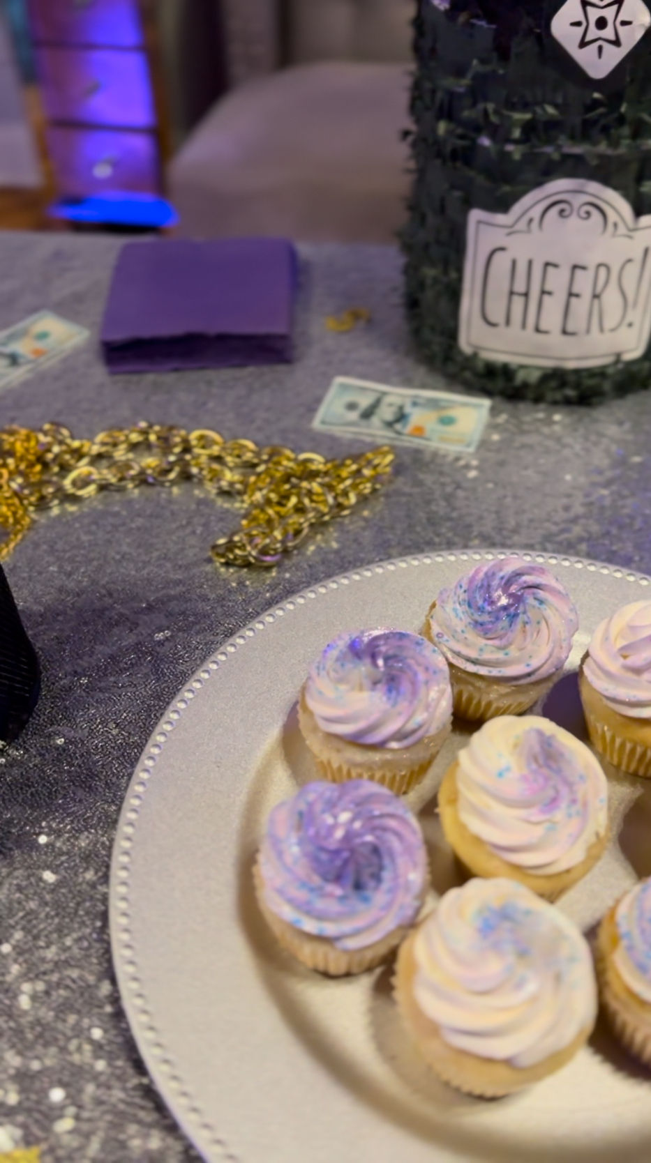 Mini cupcakes with pastel purple swirls on a silver platter at a glittery party table with gold chain, purple napkins and a jar labeled "Cheers!"