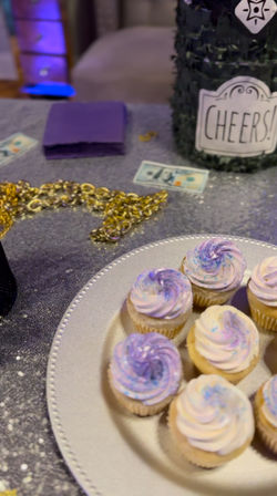 Mini cupcakes with pastel purple swirls on a silver platter at a glittery party table with gold chain, purple napkins and a jar labeled "Cheers!"