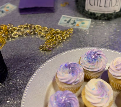Mini cupcakes with pastel purple swirls on a silver platter at a glittery party table with gold chain, purple napkins and a jar labeled "Cheers!"