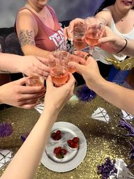 Several people clinking plastic wine glasses filled with rosé over a glittery gold party table with a plate of strawberries and purple decorations