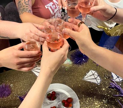 Several people clinking plastic wine glasses filled with rosé over a glittery gold party table with a plate of strawberries and purple decorations