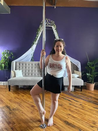 Person wearing a 'BRIDE' tank top poses on a chrome dance pole in a loft-style pole studio with a purple accent wall, tufted sofa with canopy, hardwood floors, and potted plants.