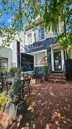 Blue two-story historic inn with cream shutters and hanging sign, shaded brick courtyard patio with black metal tables and chairs, potted plants and leafy trees on a sunny downtown street