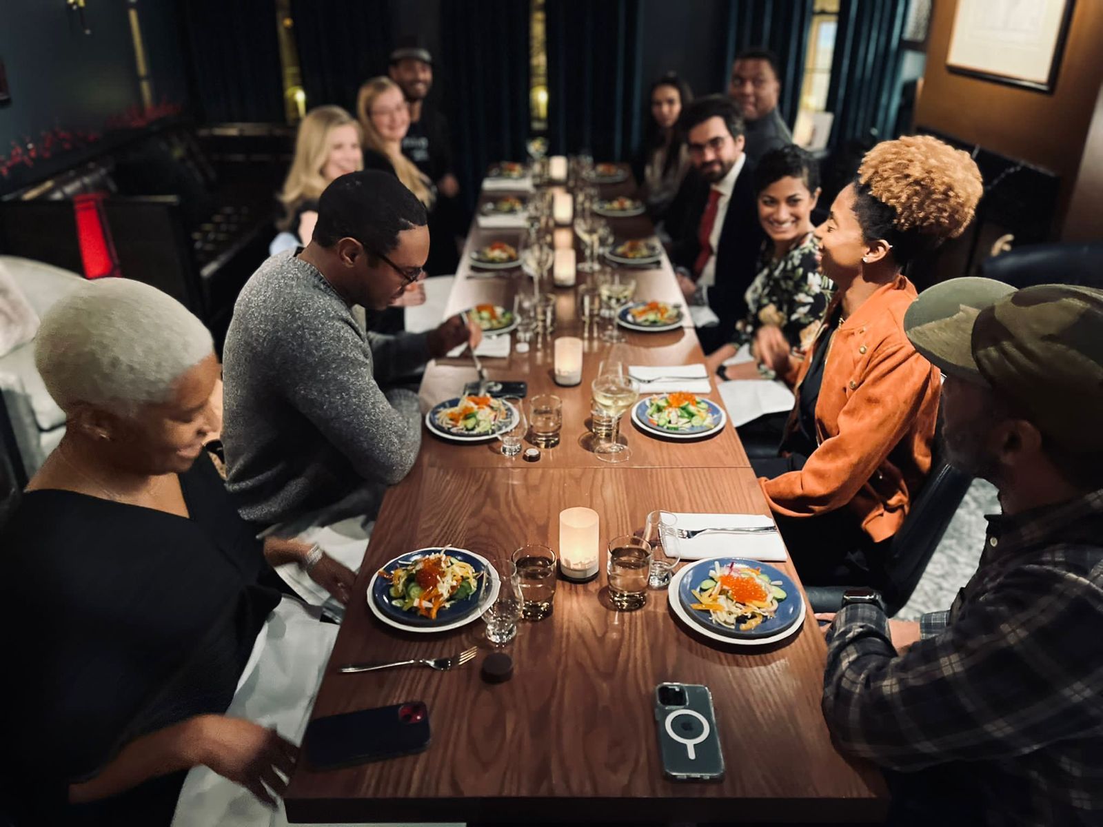 Candlelit dinner at a long communal restaurant table — a diverse group of diners chatting and laughing over plated salads, glasses of wine, and warm ambient lighting.
