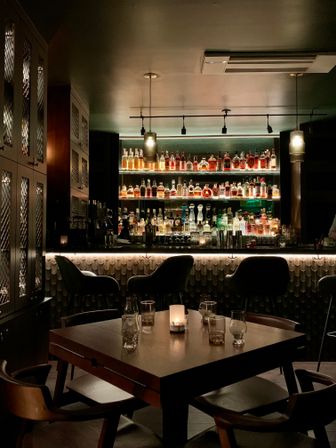 Moody candlelit cocktail bar interior with a wooden table, backlit liquor shelves and curved leather bar stools — cozy speakeasy vibe.