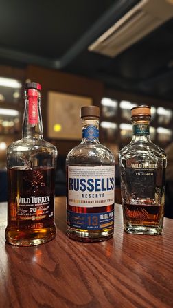 Three bourbon bottles — Wild Turkey, Russell's Reserve 13-year, and Wild Turkey Master's Keep — lined up on a wooden bar counter with blurred shelves and warm ambient lighting.