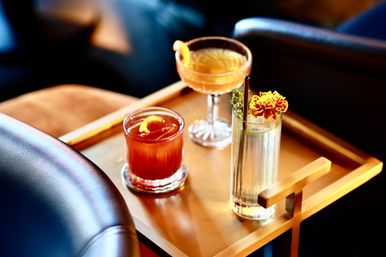 Three craft cocktails on a brass tray in a cozy lounge: a red whiskey cocktail with orange twist in a rocks glass, a pale amber coupe with lemon peel, and a tall floral highball garnished with a marigold and herb.