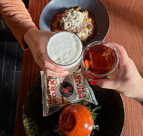 Two hands clinking drinks over a wooden restaurant table — one frothy white cocktail and one amber cocktail with a large ice cube and orange peel — beside a brioche burger bun, a packaged bag of potato chips, and a bowl of cheesy ragu-style pasta.