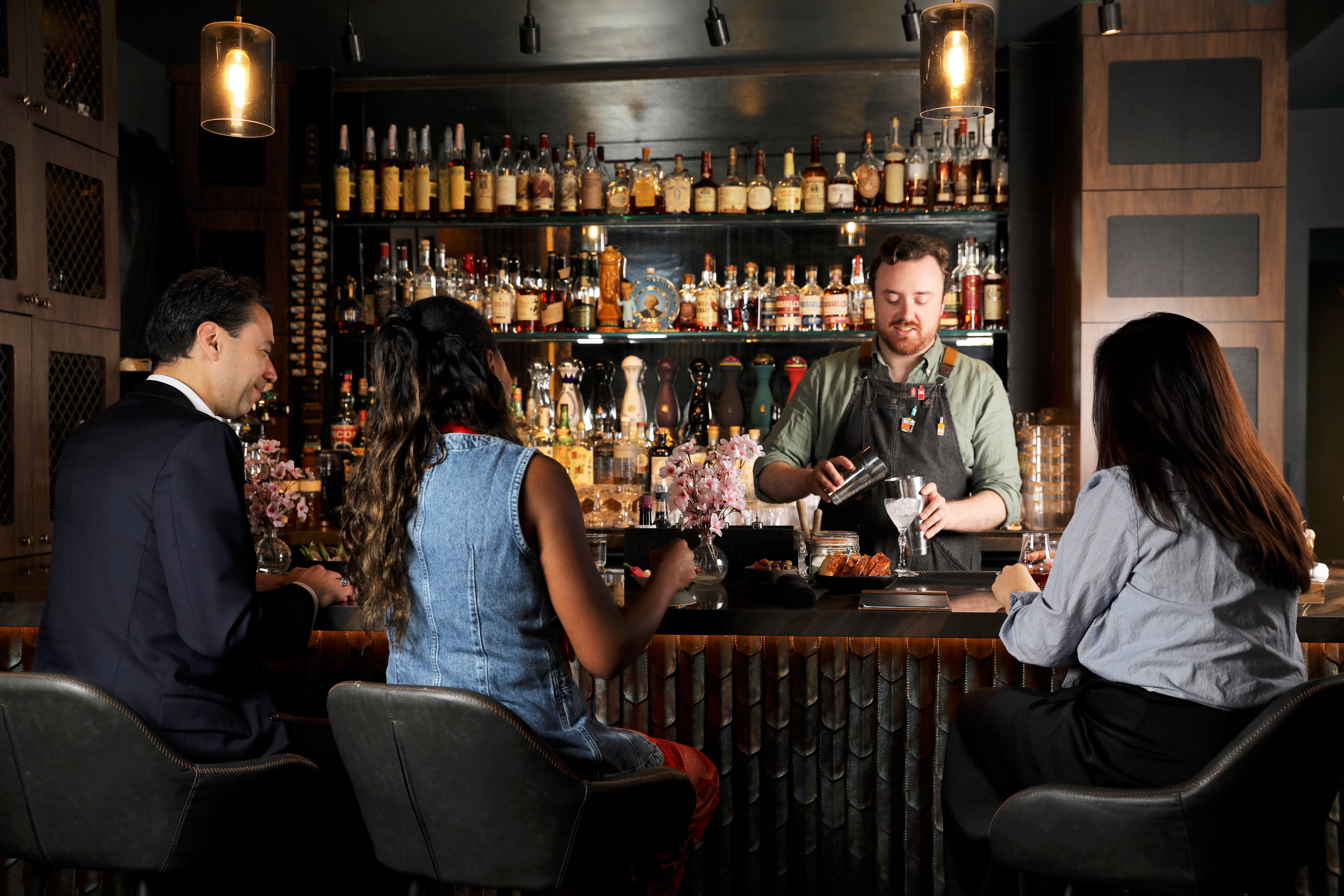 Upscale dimly lit cocktail bar — bartender pours a cocktail from a shaker while three patrons sit on barstools at a polished wood bar beneath warm pendant lights and shelves of spirits.