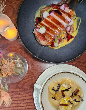 Gourmet seared pork chop sliced over grains with pickled radishes, roasted red peppers and creamy corn sauce in a black bowl, paired with dumplings in a clear corn broth topped with charred scallions, photographed on a wooden table with candlelight.