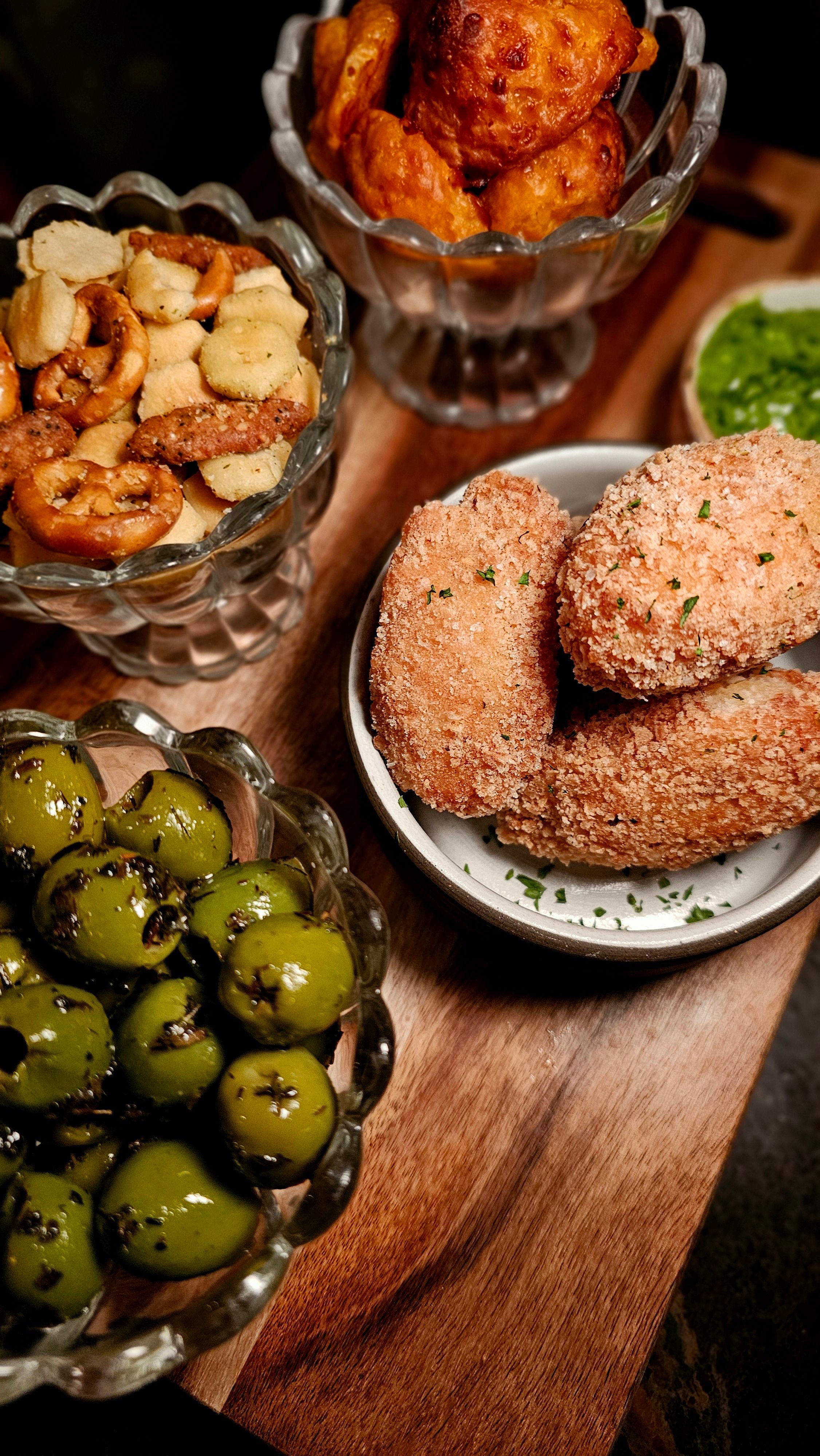 Crunchy appetizer board on a wooden serving board: golden breaded croquettes, glossy marinated green olives, mixed pretzels and crackers, and fried fritters with a green herb dip.