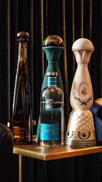 Three tall, decorative tequila bottles — a slim amber bottle, a clear bottle with a blue label, and a white ceramic decanter — arranged on a brass side table against dark velvet curtains.