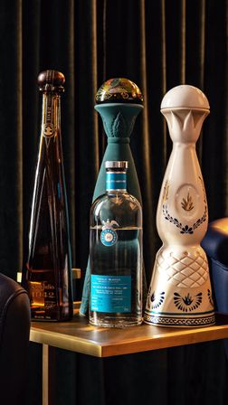 Three tall, decorative tequila bottles — a slim amber bottle, a clear bottle with a blue label, and a white ceramic decanter — arranged on a brass side table against dark velvet curtains.