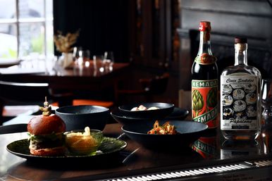 Dim, cozy restaurant table with a gourmet burger and fries on a green plate, several black bowls of small plates, and two decorative liquor bottles on a reflective bar surface.