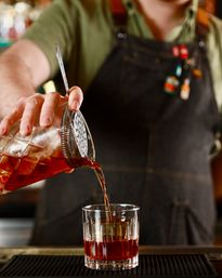 Close-up of a bartender in an apron pouring an amber craft cocktail from a mixing glass through a Hawthorne strainer into a lowball glass on a bar mat.