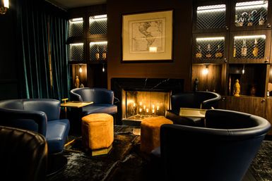 Cozy speakeasy-style lounge with a glowing fireplace, navy leather club chairs, amber suede ottomans, backlit wooden liquor cabinets, velvet curtains and a framed vintage map.