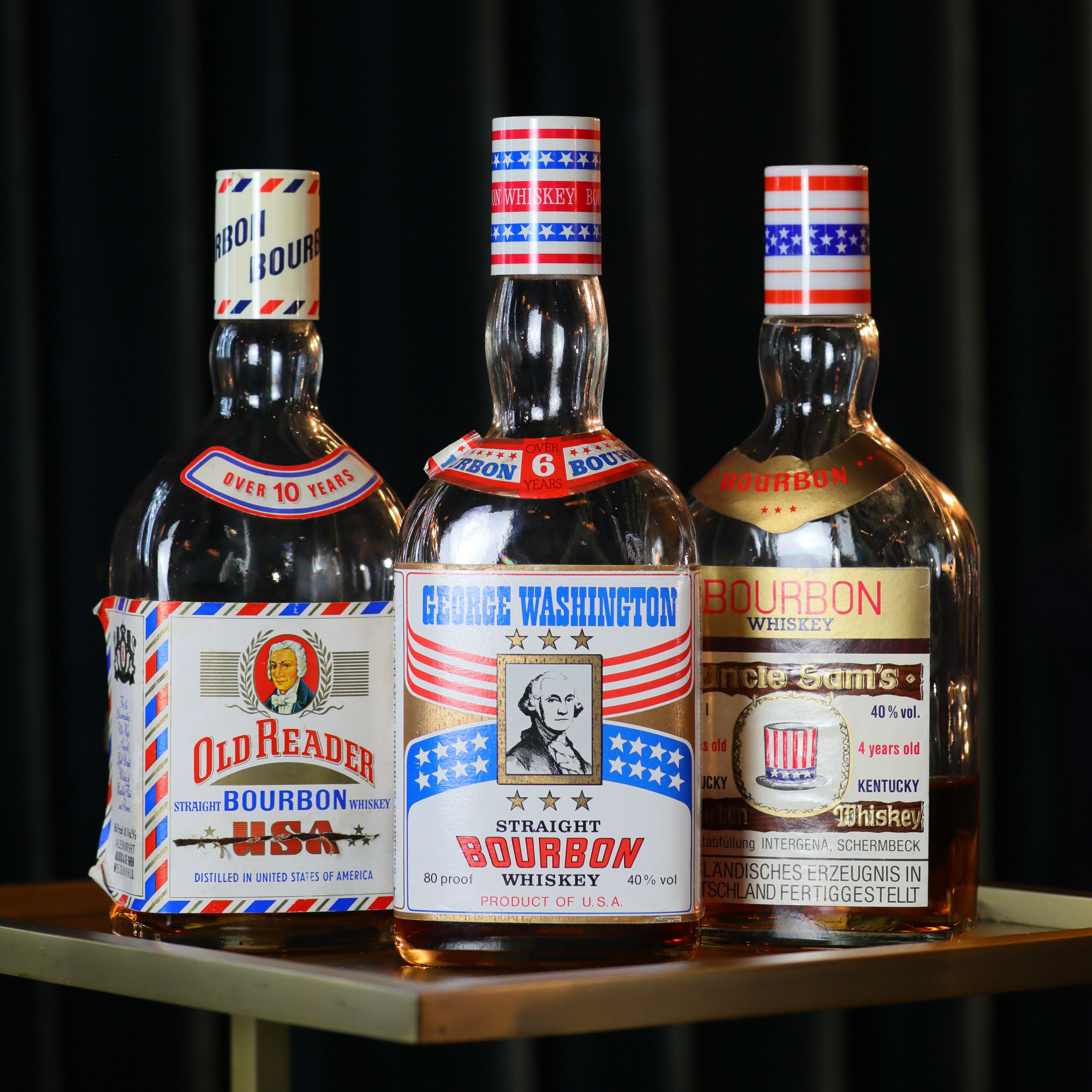 Three USA-themed bourbon whiskey bottles with red, white and blue patriotic labels and a vintage portrait on the center bottle, displayed on a small bar table