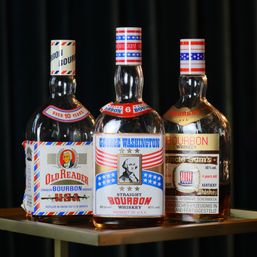 Three USA-themed bourbon whiskey bottles with red, white and blue patriotic labels and a vintage portrait on the center bottle, displayed on a small bar table