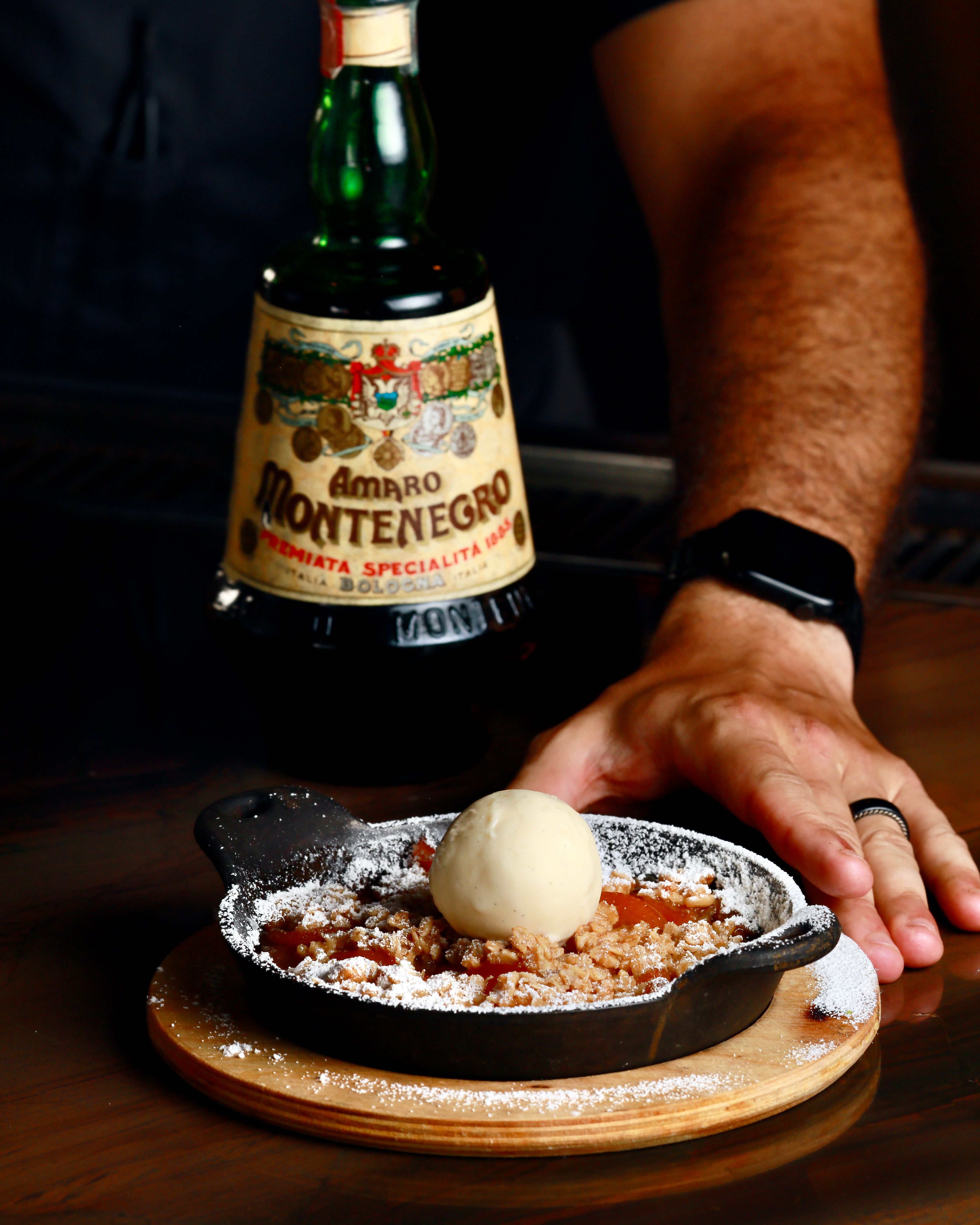 Warm cast-iron fruit crumble dusted with powdered sugar and topped with a vanilla ice cream scoop, served on a wooden board on a bar counter next to a green bottle of Italian amaro liqueur and a hand wearing a smartwatch.