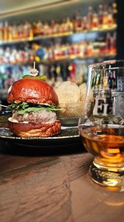 Gourmet cheeseburger on a glossy pretzel bun topped with arugula and a pickle, served with a bowl of potato chips and a whiskey glass on a wooden bar counter with blurred liquor shelves in the background.