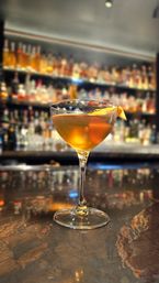 Amber cocktail in a coupe glass with an orange twist on a polished marble bar top, blurred backlit liquor shelves in the background for a cozy bar atmosphere