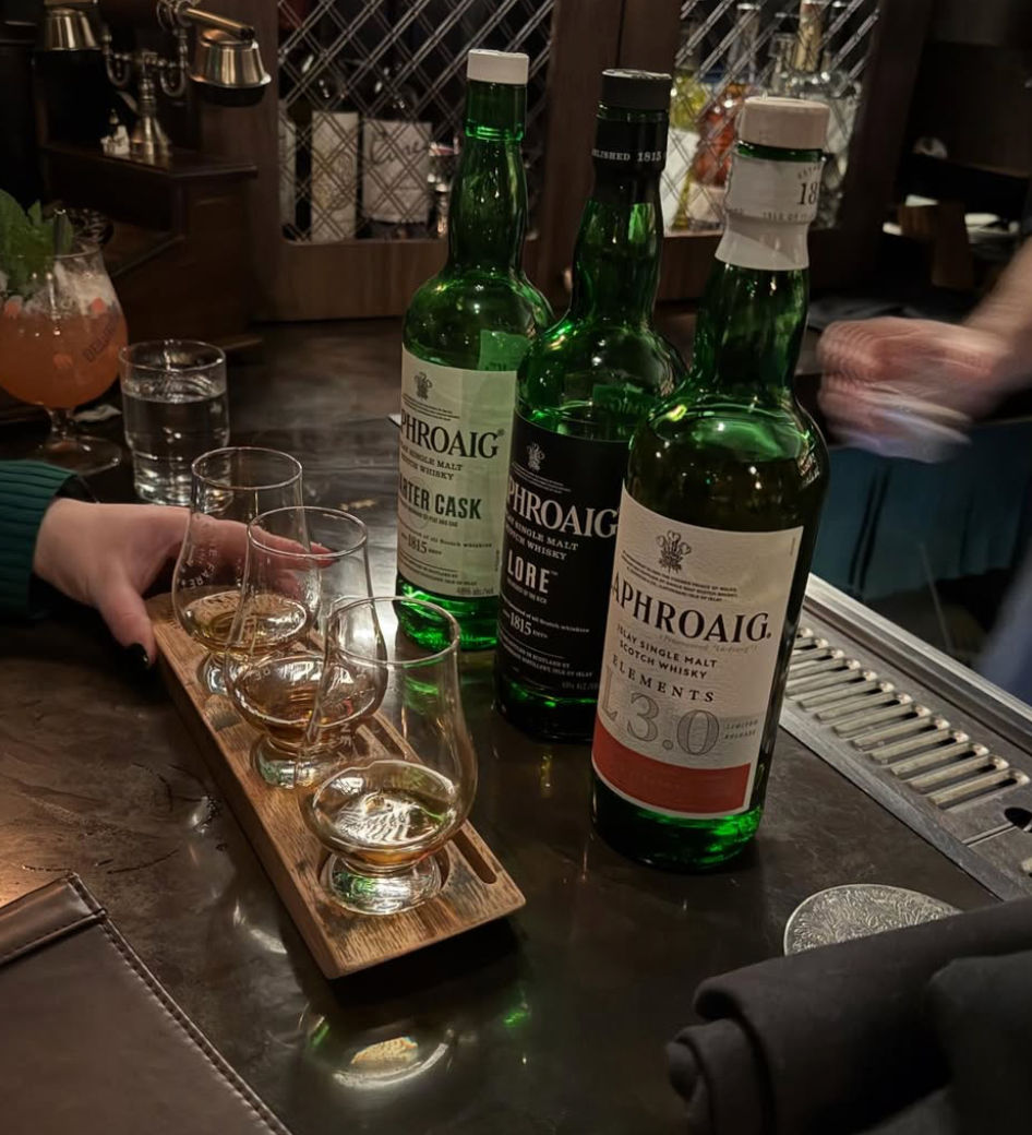 Wooden whiskey tasting flight with four tasting glasses and three green Scotch bottles arranged on a dimly lit bar counter, ready for a tasting.
