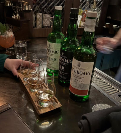 Wooden whiskey tasting flight with four tasting glasses and three green Scotch bottles arranged on a dimly lit bar counter, ready for a tasting.