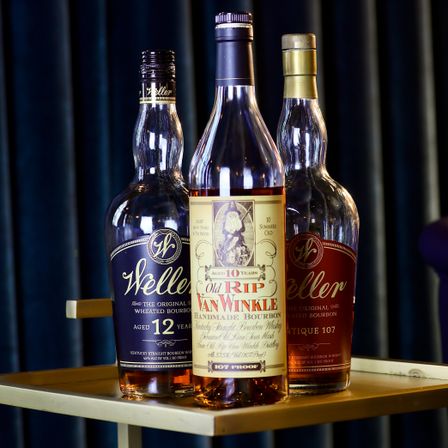 Three premium Kentucky bourbon bottles (Old Rip Van Winkle, Weller) on a gold bar cart tray, amber liquid and dark curtain backdrop