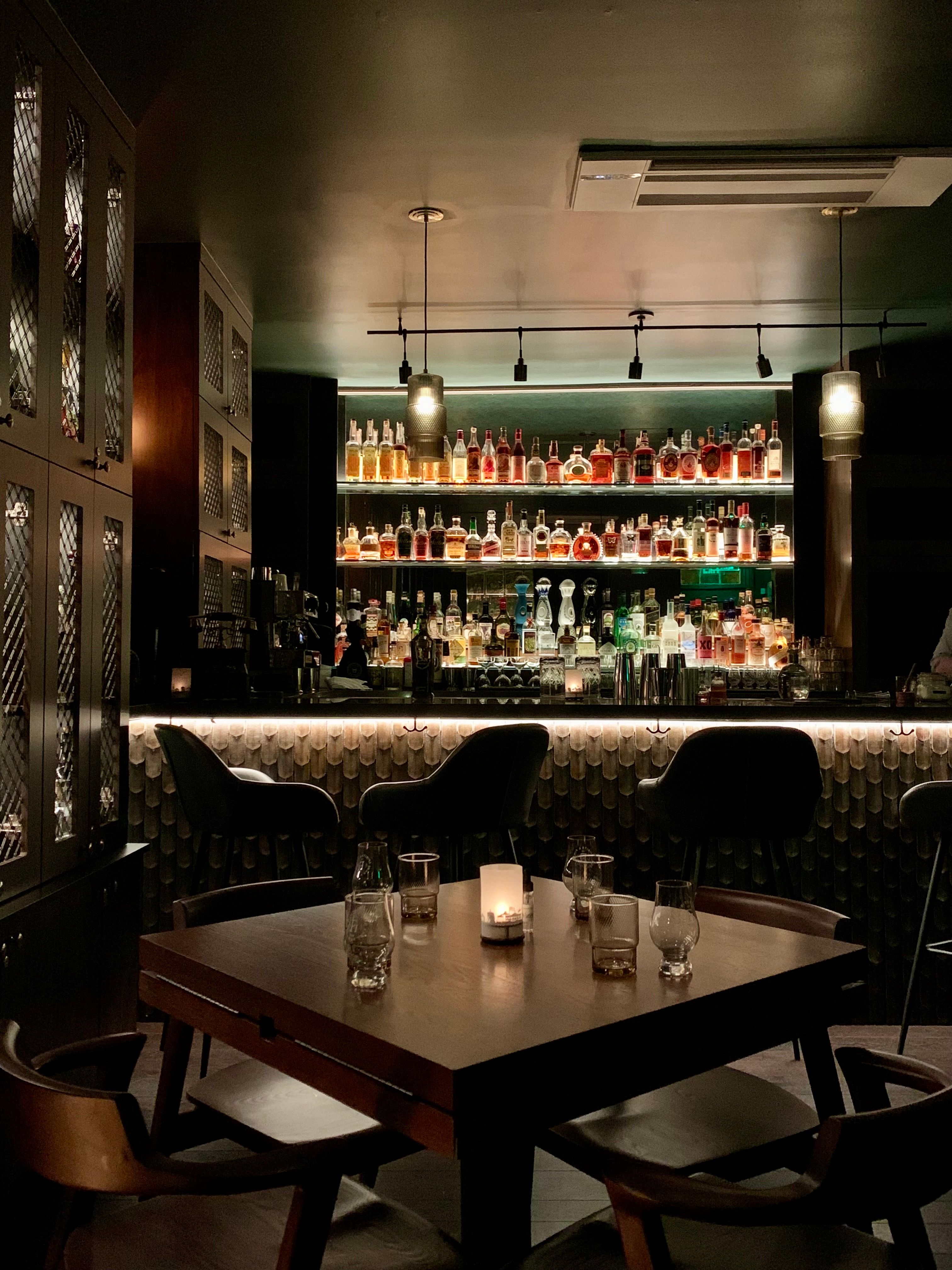 Moody, dimly lit cocktail bar interior with backlit liquor shelves, leather bar stools and a wooden table with a lit candle and empty glasses — cozy speakeasy vibe.