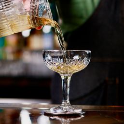 Close-up of a bartender pouring a golden cocktail through a spring strainer from a mixing glass into a cut-crystal coupe on a polished bar.