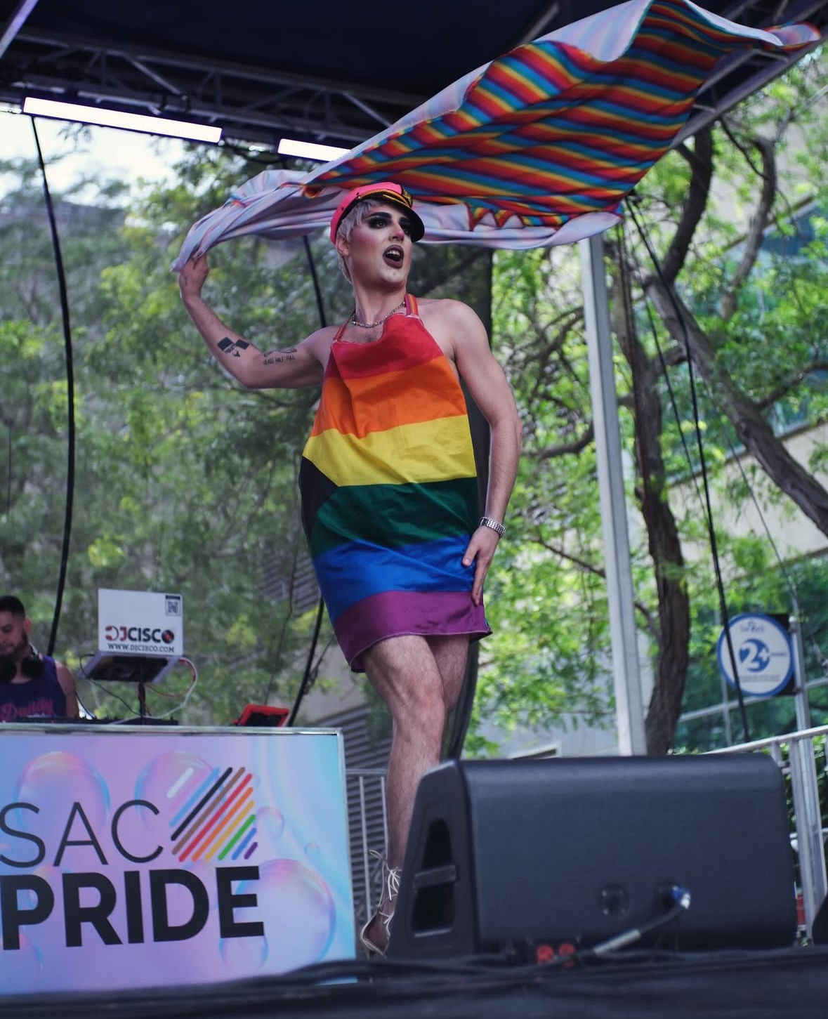 Drag performer in a rainbow-striped dress and captain cap waving a giant rainbow flag onstage at a Sacramento Pride festival with trees in the background.