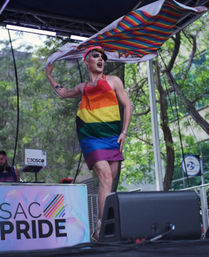 Drag performer in a rainbow-striped dress and captain cap waving a giant rainbow flag onstage at a Sacramento Pride festival with trees in the background.