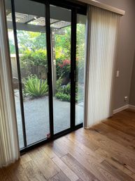 Cozy living-room sliding glass patio door with vertical blinds, oak hardwood floor in foreground, and a shaded backyard patio with ferns, shrubs, and a pergola.