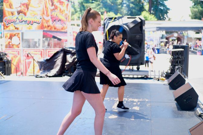 Two performers in black skater skirts dancing and singing on an outdoor festival stage by large speakers, with carnival food booths and crowds in the sunny background.